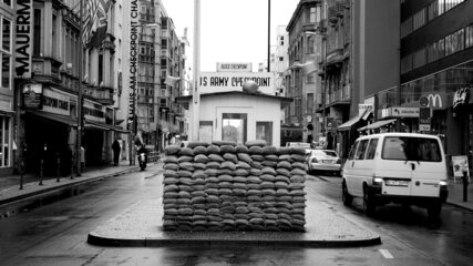 Ein gepackter Sandbags-Stapel steht auf einer Straße, umgeben von Gebäude. Im Hintergrund ist ein Kontrollpunkt sichtbar, der als "US Army Checkpoint" gekennzeichnet ist. Die Szene vermittelt ein Gefühl von historischer Bedeutung und erinnert an frühere Konflikte.
