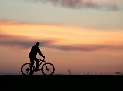 Ein Silhouettenbild eines Mannes, der ein Fahrrad hält, während er vor einem bunten Sonnenuntergang steht. Die Farben des Himmels variieren von Orange zu Lila, wodurch eine ruhige Atmosphäre entsteht.
