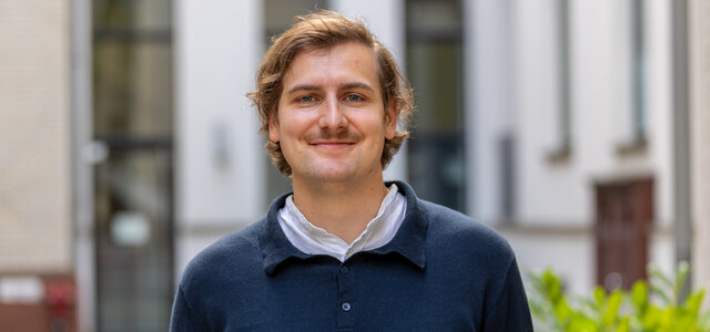 Ein junger Mann mit blonden Haaren und einem Schnurrbart trägt ein dunkelblaues Oberteil. Er steht lächelnd in einem urbanen Umfeld mit Gebäuden im Hintergrund. Die Atmosphäre wirkt freundlich und einladend.