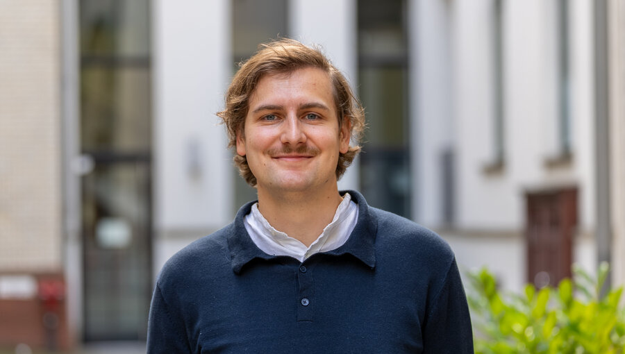 Ein junger Mann mit blonden Haaren und einem Schnurrbart trägt ein dunkelblaues Oberteil. Er steht lächelnd in einem urbanen Umfeld mit Gebäuden im Hintergrund. Die Atmosphäre wirkt freundlich und einladend.