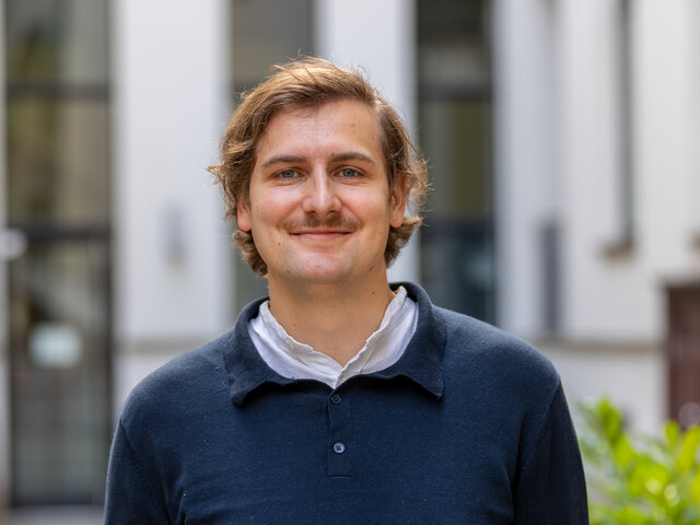 Ein junger Mann mit blonden Haaren und einem Schnurrbart trägt ein dunkelblaues Oberteil. Er steht lächelnd in einem urbanen Umfeld mit Gebäuden im Hintergrund. Die Atmosphäre wirkt freundlich und einladend.