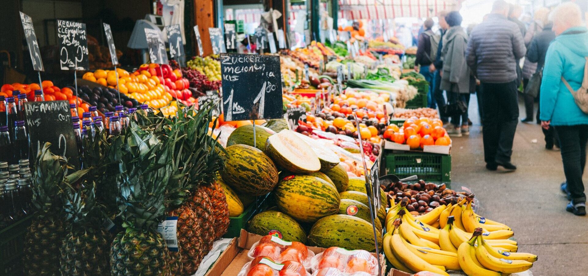 Ein lebhafter Markt mit verschiedenem Obst und Gemüse. Im Vordergrund sind Ananas, Melonen, Bananen und verpackte Lebensmittel zu sehen. Händler präsentieren ihre Waren auf Tischplatten, während Kunden durch die Gänge schlendern und die Farben und Vielfalt der Produkte genießen.