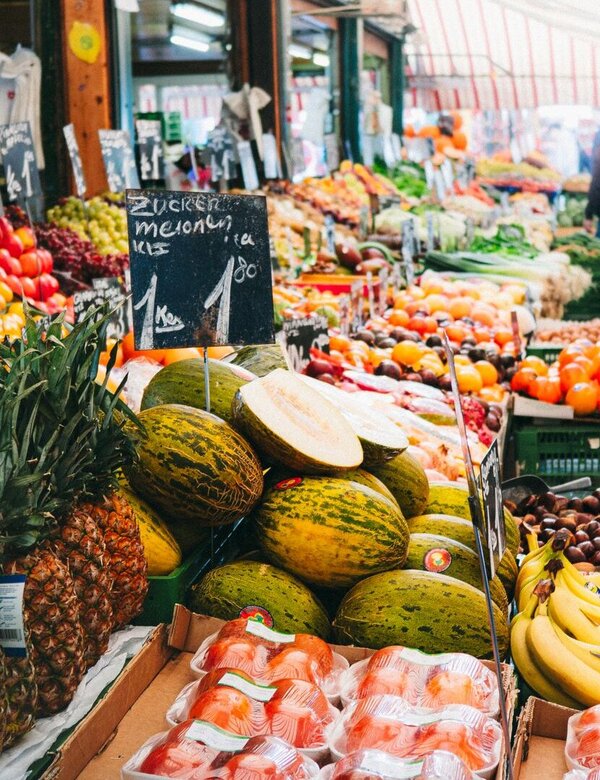 Ein lebhafter Markt mit verschiedenem Obst und Gemüse. Im Vordergrund sind Ananas, Melonen, Bananen und verpackte Lebensmittel zu sehen. Händler präsentieren ihre Waren auf Tischplatten, während Kunden durch die Gänge schlendern und die Farben und Vielfalt der Produkte genießen.