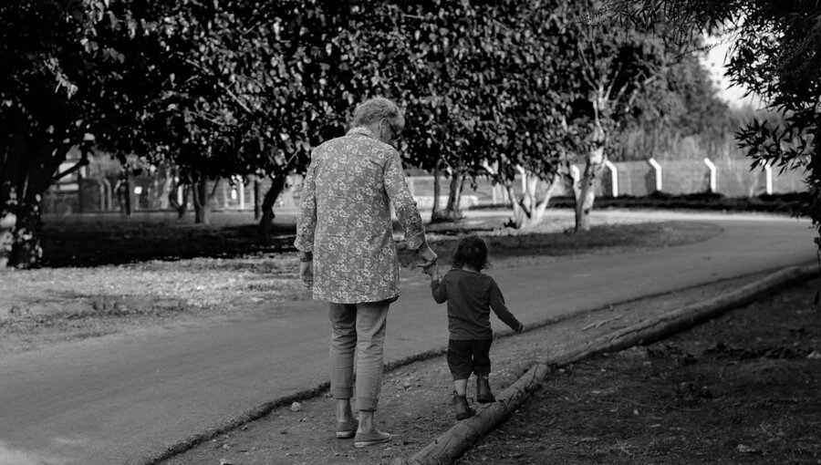 Eine ältere Frau und ein kleines Kind gehen Hand in Hand auf einem Weg durch einen Park. Um sie herum sind Bäume und die Umgebung wirkt friedlich und einladend.