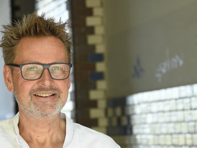 Ein lächelnder Mann mit kurzen, strubbeligen Haaren und einer Brille steht in einem hellen Raum. Er trägt ein weißes Hemd und wirkt freundlich und zugänglich. Im Hintergrund sind farbige Fliesen sichtbar, die dem Raum eine künstlerische Note verleihen.