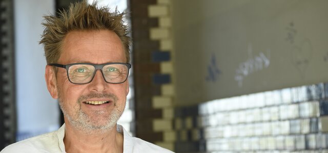 Ein lächelnder Mann mit kurzen, strubbeligen Haaren und einer Brille steht in einem hellen Raum. Er trägt ein weißes Hemd und wirkt freundlich und zugänglich. Im Hintergrund sind farbige Fliesen sichtbar, die dem Raum eine künstlerische Note verleihen.