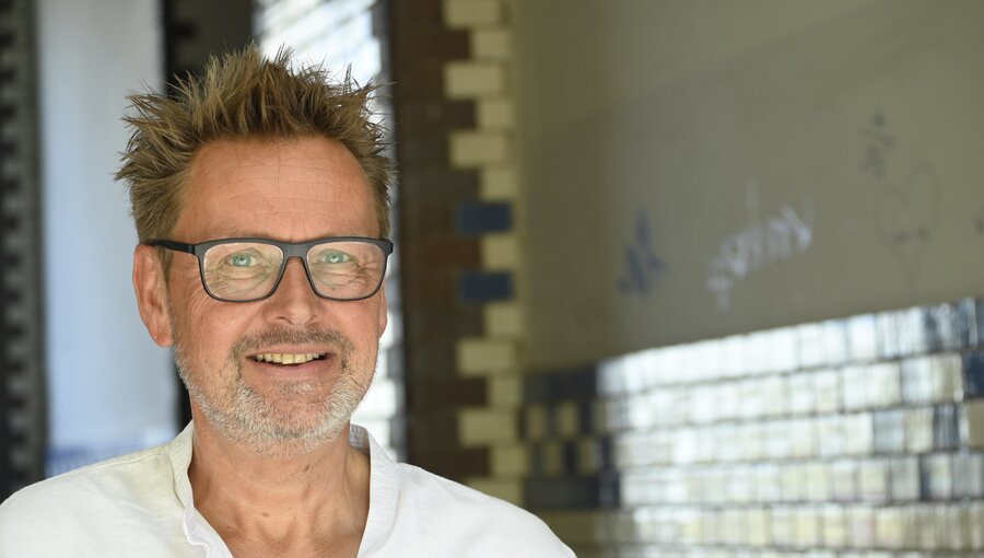 Ein lächelnder Mann mit kurzen, strubbeligen Haaren und einer Brille steht in einem hellen Raum. Er trägt ein weißes Hemd und wirkt freundlich und zugänglich. Im Hintergrund sind farbige Fliesen sichtbar, die dem Raum eine künstlerische Note verleihen.