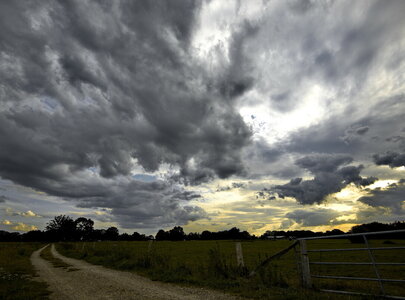Ein unebener Weg führt durch eine weite Wiese unter einem dramatischen Himmel, der mit dunklen, dichter werdenden Wolken bedeckt ist. Am Horizont ist ein sanfter Sonnenuntergang zu sehen, der die Landschaft in warmen Farben erhellt.