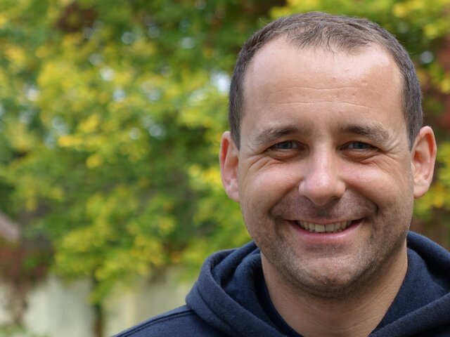 Ein lächelnder Mann trägt einen dunkelblauen Hoodie. Er steht vor einer unscharfen grünen Hintergrundpflanze, die Herbstlaub zeigt. Der Mann hat kurzes, dunkles Haar und strahlt Freundlichkeit aus.