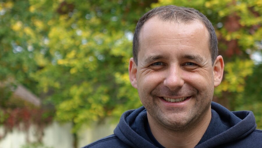 Ein lächelnder Mann trägt einen dunkelblauen Hoodie. Er steht vor einer unscharfen grünen Hintergrundpflanze, die Herbstlaub zeigt. Der Mann hat kurzes, dunkles Haar und strahlt Freundlichkeit aus.