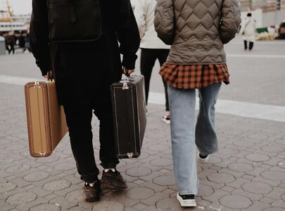 Zwei Personen gehen nebeneinander auf einem Platz, während sie jeweils einen Koffer in der Hand halten. Die Person links trägt einen schwarzen Mantel, die Person rechts hat eine grüne Jacke und trägt eine karierte Bluse unterhalb.