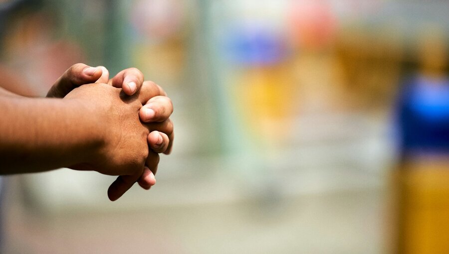 Zwei Hände sind gefaltet und ruhen aufeinander. Der Fokus liegt auf der Handhaltung, während der Hintergrund unscharf ist. Dies vermittelt einen Eindruck von Nachdenklichkeit oder Geduld.