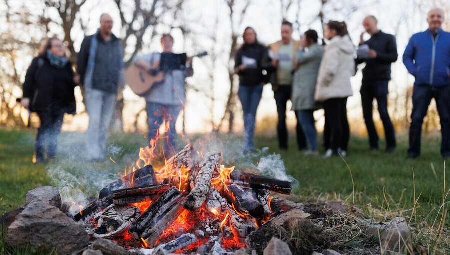 Ein knisterndes Lagerfeuer brennt im Vordergrund, umgeben von Steinen. Im Hintergrund stehen mehrere Personen, einige applaudieren, während eine Person mit einer Gitarre spielt. Die Szene vermittelt eine Atmosphäre von Geselligkeit und Feierlichkeit in der Natur.