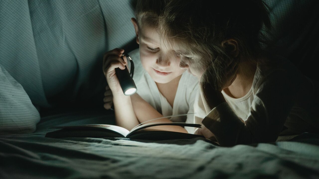 Zwei Kinder sitzen unter einer Decke und lesen ein Buch. Eines der Kinder hält eine Taschenlampe, die das Buch beleuchtet. Die Umgebung ist schwach beleuchtet, und die Gesichter der Kinder zeigen Interesse und Konzentration beim Lesen.