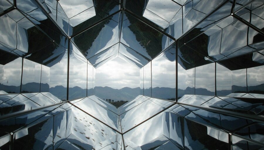 Das Bild zeigt eine geometrische Struktur aus verspiegelten Flächen, die den Himmel und die umliegenden Berge reflektiert. Die Anordnung der Spiegel erzeugt eine kaleidoskopartige Wirkung, die das Licht und die Landschaft auf interessante Weise verzerrt und vervielfältigt.