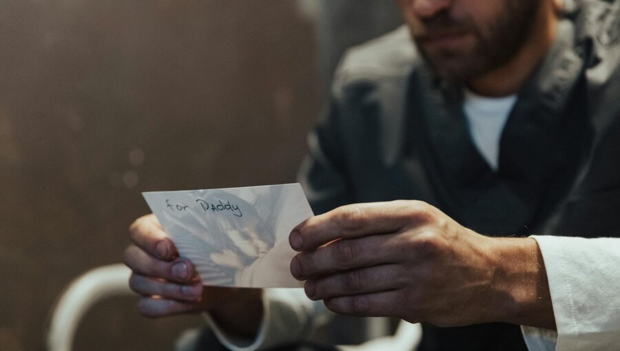 Ein Mann sitzt und hält eine Karte in den Händen. Auf der Karte steht handschriftlich "for Daddy". Der Ausdruck auf dem Gesicht des Mannes zeigt Nachdenklichkeit oder Emotion.