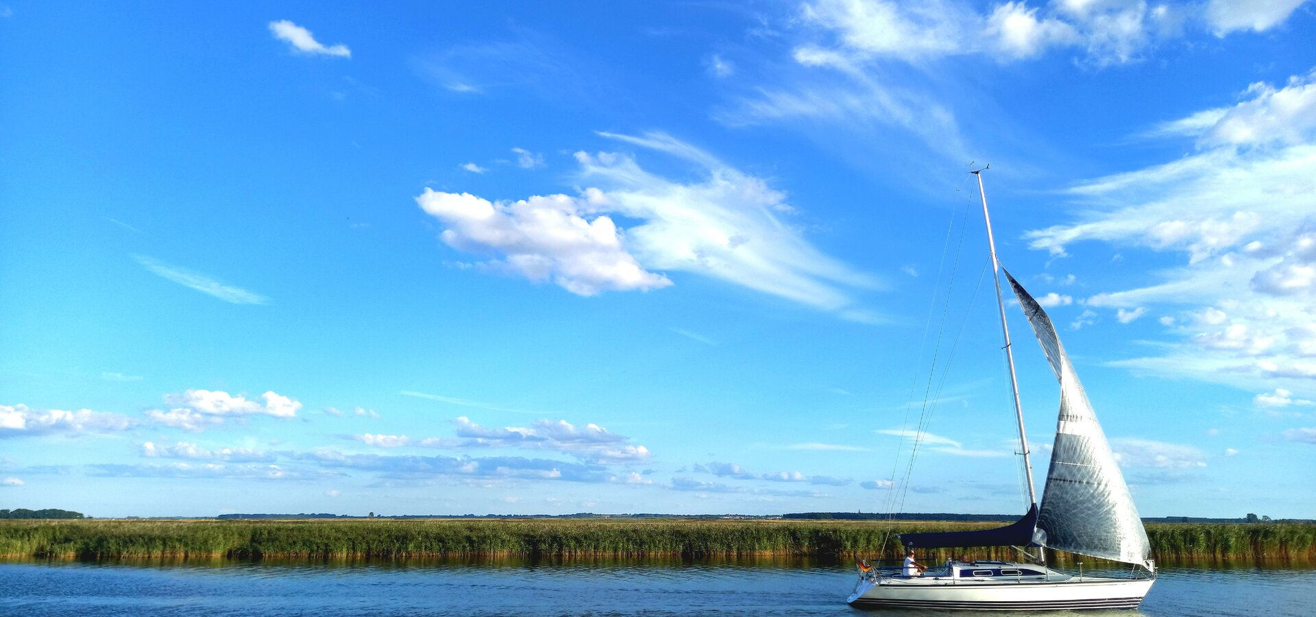 Ein Segelboot fährt ruhig auf einem Gewässer, umgeben von grünen Uferpflanzen. Der Himmel ist blau mit weißen Wolken, die sanft dahin ziehen. Die Szenerie vermittelt ein Gefühl von Frieden und Freiheit in der Natur.
