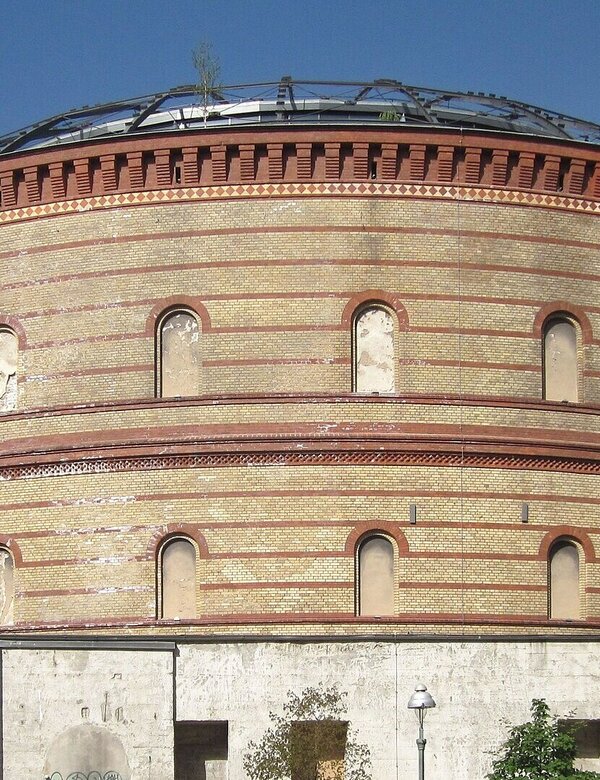 Ein historisches, rundes Gebäude mit einer auffälligen Ziegelmauer. Das Dach ist gewölbt und teilweise begrünt. Vor dem Eingang befinden sich Menschen und Autos. Das Gebäude scheint eine kulturelle oder öffentliche Nutzung zu haben.