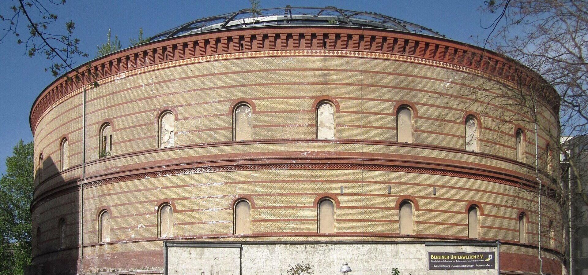 Ein historisches, rundes Gebäude mit einer auffälligen Ziegelmauer. Das Dach ist gewölbt und teilweise begrünt. Vor dem Eingang befinden sich Menschen und Autos. Das Gebäude scheint eine kulturelle oder öffentliche Nutzung zu haben.
