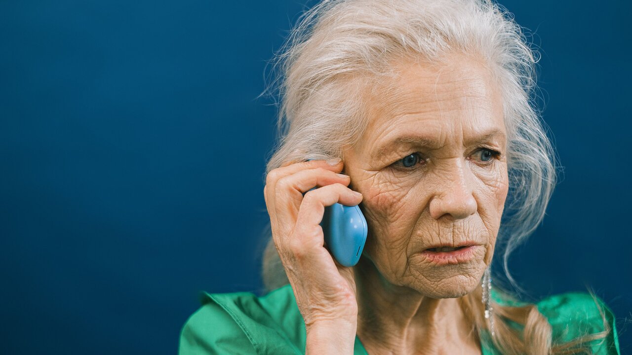 Eine ältere Frau mit silbernem Haar trägt ein grünes Oberteil und hält ein blaues Telefon am Ohr. Sie schaut besorgt und konzentriert aus, während sie spricht. Der Hintergrund ist in einem neutralen Blau gehalten.