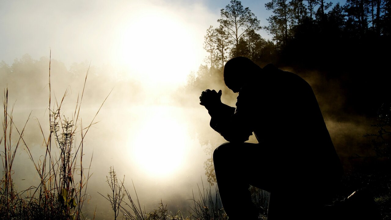 Eine Silhouette einer Person, die vor einem nebligen Gewässer kniet. Die Sonne scheint schwach hinter der Person und reflektiert im Wasser. Umgeben von Bäumen und Gras, vermittelt die Szene eine ruhige und nachdenkliche Atmosphäre.