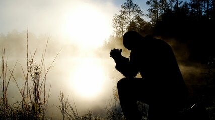 Eine Silhouette einer Person, die vor einem nebligen Gewässer kniet. Die Sonne scheint schwach hinter der Person und reflektiert im Wasser. Umgeben von Bäumen und Gras, vermittelt die Szene eine ruhige und nachdenkliche Atmosphäre.