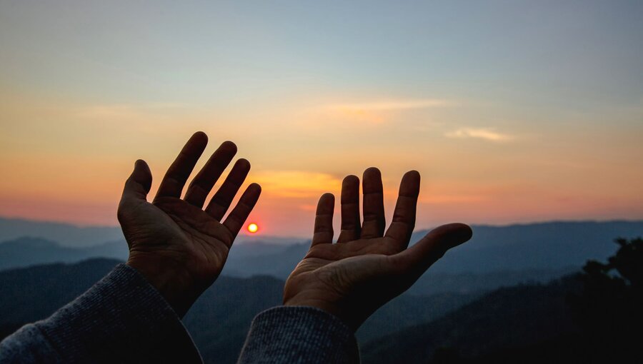 Zwei Hände erheben sich gegen einen Sonnenuntergang, während die Sonne am Horizont aufgeht. Die Silhouette der Berge bildet den Hintergrund, und die Farben des Himmels reichen von orange bis blau, was eine friedliche und ergreifende Stimmung vermittelt.