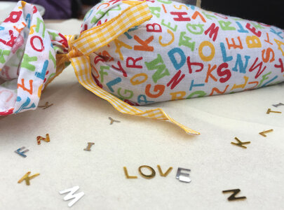 Ein buntes, mit Buchstaben bedrucktes Tuch, das wie ein Bonbon aussieht, liegt auf einem weißen Tisch. Um das Tuch herum sind silberne und goldene Buchstaben verteilt, die das Wort „LOVE“ bilden.