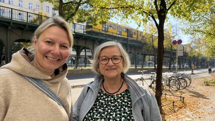 Zwei Frauen lächeln in einem urbanen Umfeld. Im Hintergrund ist eine Unterführung mit einer U-Bahn und bunten Herbstblättern sichtbar. Die erste Frau trägt einen hellen Pullover, die zweite eine Jacke und Brille.