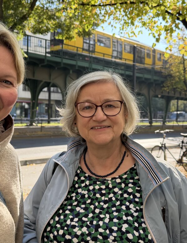 Zwei Frauen lächeln in einem urbanen Umfeld. Im Hintergrund ist eine Unterführung mit einer U-Bahn und bunten Herbstblättern sichtbar. Die erste Frau trägt einen hellen Pullover, die zweite eine Jacke und Brille.