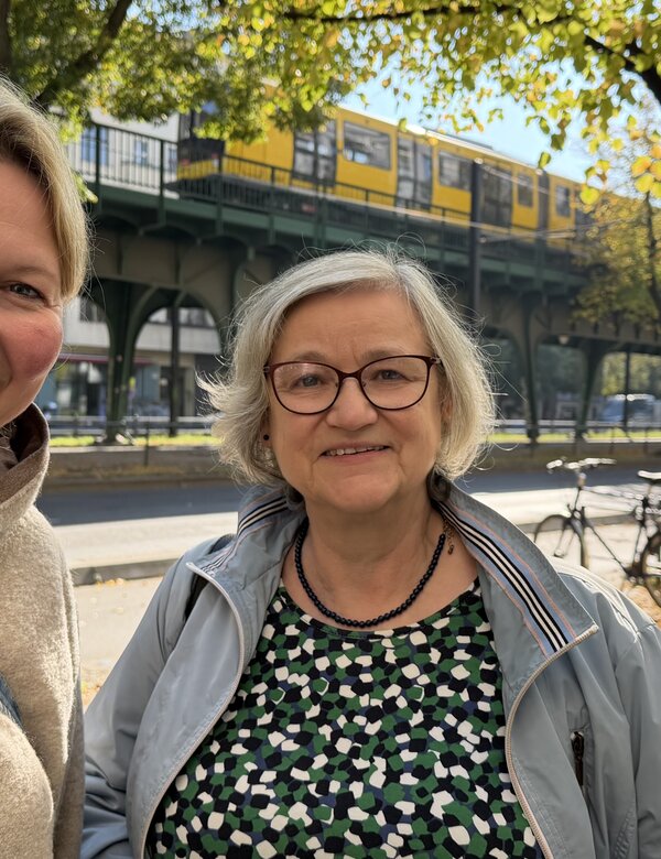 Zwei Frauen lächeln in einem urbanen Umfeld. Im Hintergrund ist eine Unterführung mit einer U-Bahn und bunten Herbstblättern sichtbar. Die erste Frau trägt einen hellen Pullover, die zweite eine Jacke und Brille.