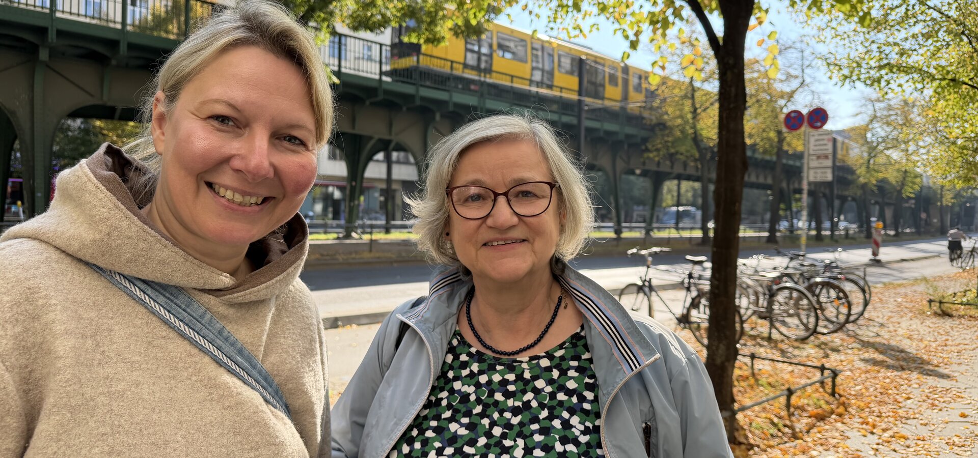 Zwei Frauen lächeln in einem urbanen Umfeld. Im Hintergrund ist eine Unterführung mit einer U-Bahn und bunten Herbstblättern sichtbar. Die erste Frau trägt einen hellen Pullover, die zweite eine Jacke und Brille.