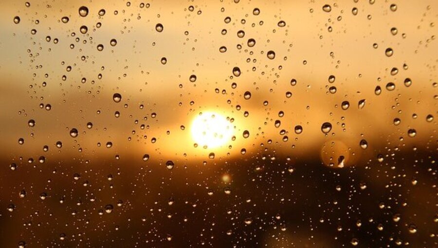 Wassertropfen liegen auf einer Fensterscheibe, während die Sonne am Horizont untergeht. Die warmen Farben des Sonnenuntergangs spiegeln sich in den Tropfen wider und schaffen eine ruhige, nostalgische Atmosphäre.