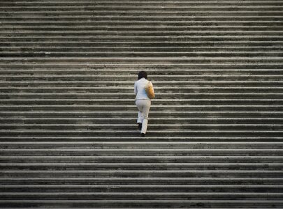 Eine Person in weißer Kleidung steigt eine lange Treppe empor. Die Stufen sind gleichmäßig und in einem neutralen Farbton gehalten. Der Fokus liegt auf dem aufstrebenden Charakter und der Herausforderung des Aufstiegs.
