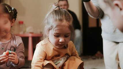 Ein kleines Mädchen mit blonden Haaren sitzt auf dem Boden und spielt mit Spielzeug. Sie trägt ein orangefarbenes Shirt und ist konzentriert bei der Sache. Im Hintergrund sind andere Kinder und Erwachsene zu sehen, die ebenfalls am Spiel beteiligt sind.