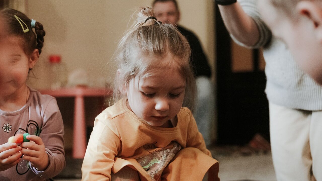 Ein kleines Mädchen mit blonden Haaren sitzt auf dem Boden und spielt mit Spielzeug. Sie trägt ein orangefarbenes Shirt und ist konzentriert bei der Sache. Im Hintergrund sind andere Kinder und Erwachsene zu sehen, die ebenfalls am Spiel beteiligt sind.