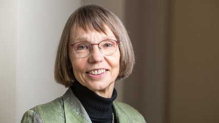 Porträt einer Frau mit kurzen, grauen Haaren, die eine grüne Jacke und einen schwarzen Rollkragenpullover trägt. Sie lächelt freundlich und trägt eine Brille. Der Hintergrund ist hell und unscharf, was den Fokus auf die Frau lenkt.