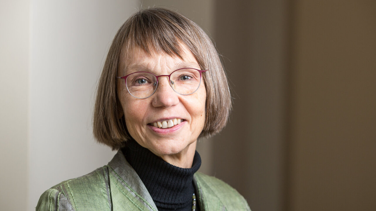 Porträt einer Frau mit kurzen, grauen Haaren, die eine grüne Jacke und einen schwarzen Rollkragenpullover trägt. Sie lächelt freundlich und trägt eine Brille. Der Hintergrund ist hell und unscharf, was den Fokus auf die Frau lenkt.