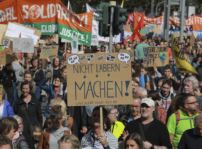 Eine große Menschenmenge nimmt an einer Demonstration teil. Viele halten Plakate mit verschiedenen Botschaften hoch. Im Vordergrund ist ein Plakat zu sehen, das den Satz "NICHT LABERN, MACHEN!" trägt. Die Stimmung ist aktiv und engagiert.