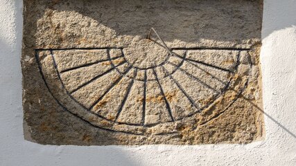 Auf einer Wand befindet sich eine Sonnenuhr aus Stein. Sie zeigt ein halbkreisförmiges Ziffernblatt mit radialen Linien, die die Stunden markieren. Ein Zeiger projiziert einen Schatten auf die Linien, der die Zeit anzeigt.