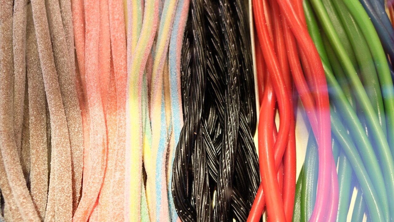 Eine Auswahl an farbigen Süßwaren in Form von langen, schlanken Streifen. Die Farben umfassen Rosa, Blau, Schwarz, Rot und Grün, die nebeneinander angeordnet sind. Die Textur der Süßigkeiten wirkt weich und gleitend.