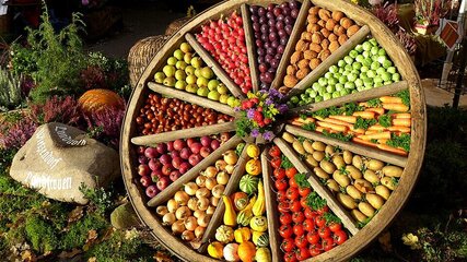 Ein großer Holzrad befindet sich im Freien, gefüllt mit verschiedenen Obst- und Gemüsesorten, die farbenfroh angeordnet sind. Die Segmente enthalten Äpfel, Birnen, Trauben, Nüsse, Kürbisse und Karotten. In der Mitte ist ein kleiner Blumenschmuck platziert.
