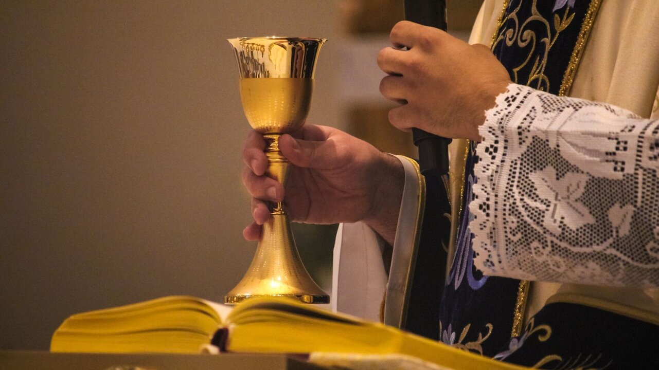 Ein Priester hält einen goldenen Kelch in der linken Hand, während er mit der rechten Hand ein offenes liturgisches Buch auf einem Tisch stützt. Der Priester trägt einen festlichen Gewand mit dekorativen Mustern.