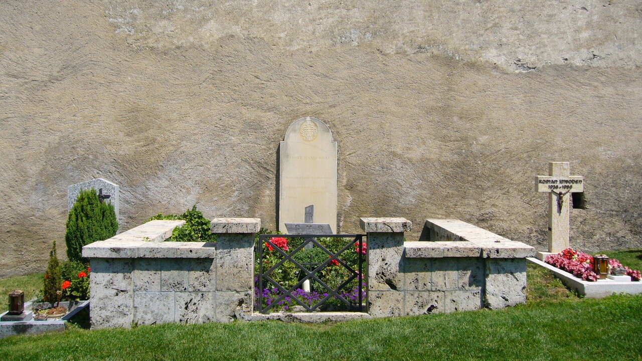 Ein ruhiger Friedhof mit Grabstätten. Im Vordergrund befindet sich eine Blumenumrandung und eine kleine Mauer. Im Hintergrund steht eine kahle Wand. Links sind schlichte Grabsteine, während rechts ein Kreuz sichtbar ist, umgeben von bunten Blumen.