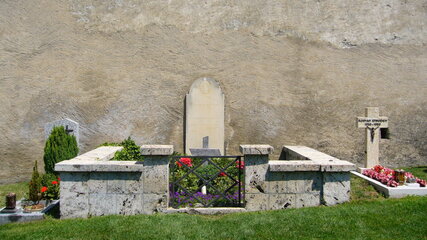 Ein ruhiger Friedhof mit Grabstätten. Im Vordergrund befindet sich eine Blumenumrandung und eine kleine Mauer. Im Hintergrund steht eine kahle Wand. Links sind schlichte Grabsteine, während rechts ein Kreuz sichtbar ist, umgeben von bunten Blumen.