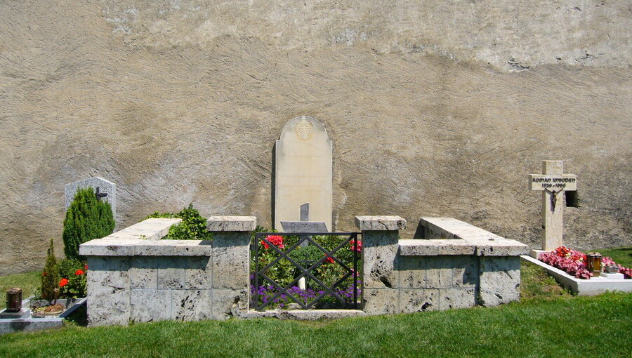 Ein ruhiger Friedhof mit Grabstätten. Im Vordergrund befindet sich eine Blumenumrandung und eine kleine Mauer. Im Hintergrund steht eine kahle Wand. Links sind schlichte Grabsteine, während rechts ein Kreuz sichtbar ist, umgeben von bunten Blumen.