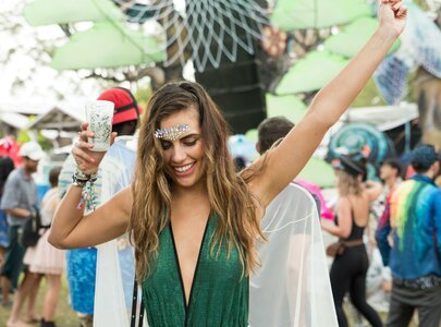 Eine fröhliche Frau tanzt in einem Festivalumfeld, trägt ein glitzerndes Outfit und hebt ein Getränk hoch. Um sie herum sind andere Festivalbesucher in bunten Kleidungsstücken, die die ausgelassene Atmosphäre des Events vermitteln.