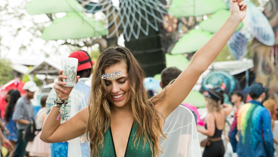 Eine fröhliche Frau tanzt in einem Festivalumfeld, trägt ein glitzerndes Outfit und hebt ein Getränk hoch. Um sie herum sind andere Festivalbesucher in bunten Kleidungsstücken, die die ausgelassene Atmosphäre des Events vermitteln.