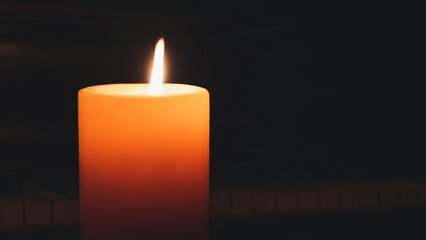 Eine brennende, orangefarbene Kerze steht auf einer dunklen Oberfläche. Die Flamme leuchtet sanft und erzeugt eine ruhige Atmosphäre. Im Hintergrund sind unscharfe Konturen zu erkennen, die an ein Klavier erinnern.