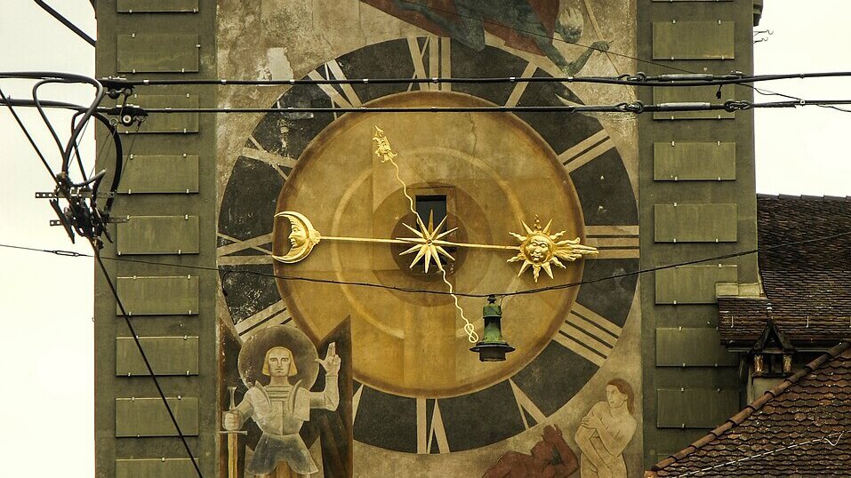 Eine große, historische Uhr ist an der Wand eines Gebäudes zu sehen. Der Ziffernblatt zeigt goldene Zeiger und ist reich verziert mit Bildern von einem Engel und einer Figur. Die Uhr ist in einem städtischen Umfeld mit Stromleitungen im Vordergrund platziert.